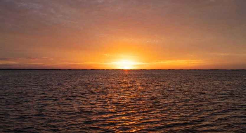 The sky is orange as the sun sets on the horizon above a body of water. 